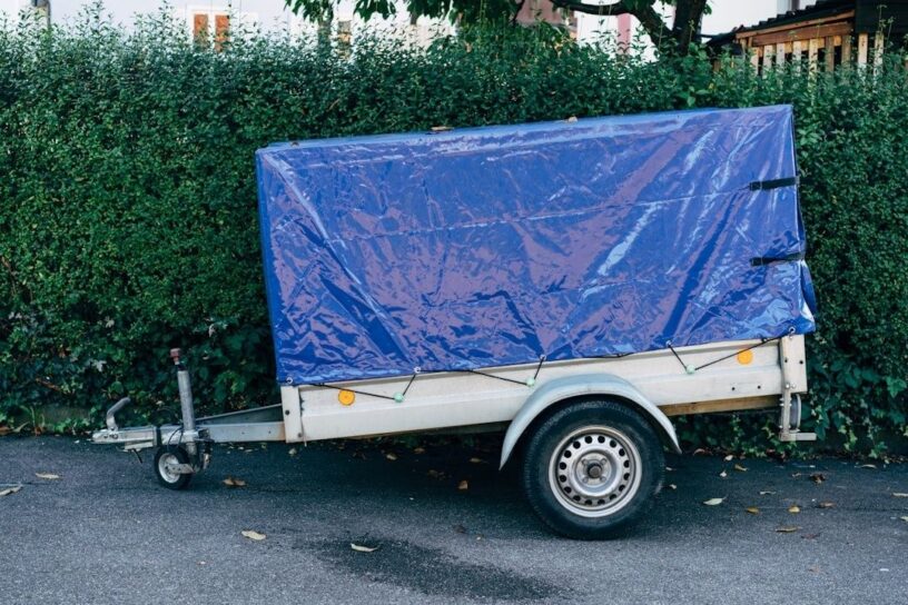 Tilhenger med blå presenning parkert ved en hekk